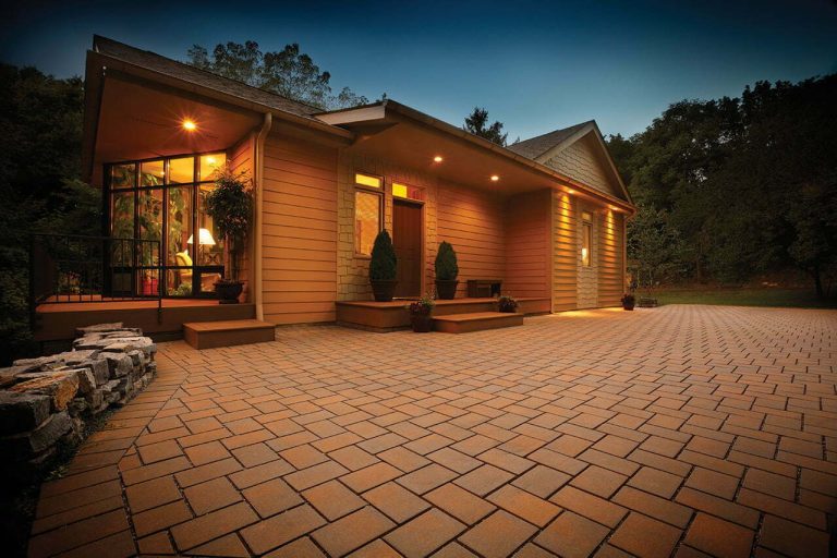 Single-story house with warm lights, large windows, and a spacious paved driveway at dusk.