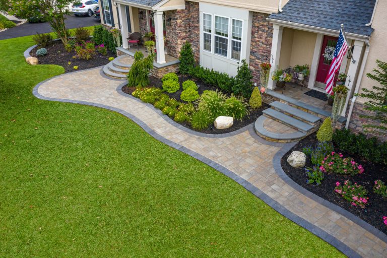 A curved stone walkway leads to a house entrance, bordered by green grass and landscaped garden beds.
