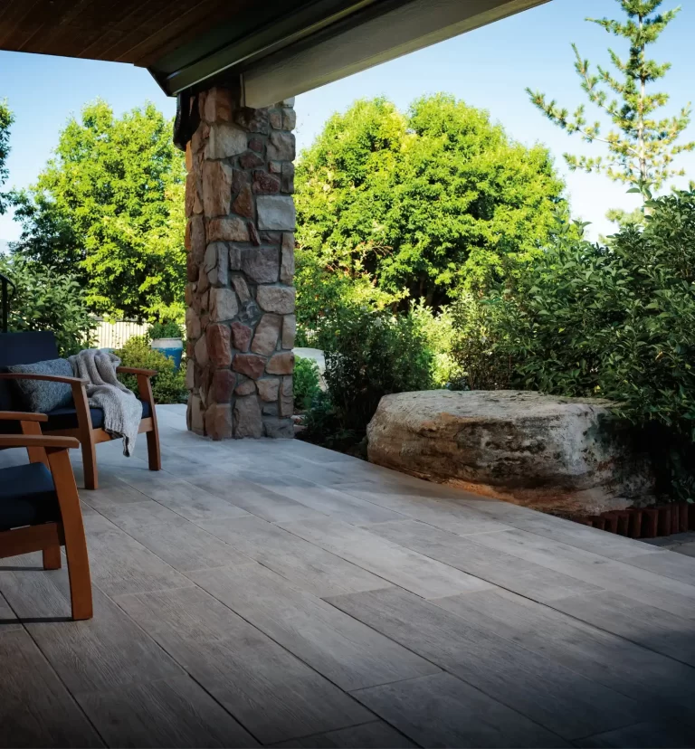 Patio with wooden chairs, stone pillar, large stone, and lush green trees under a clear blue sky.