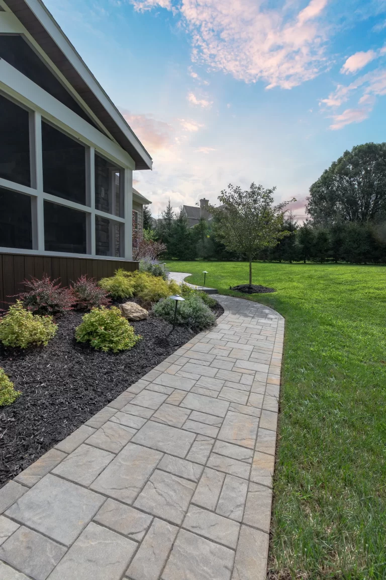 Stone walkway curves past mulched garden beds and grass beside a screened-in porch under a blue sky with clouds.