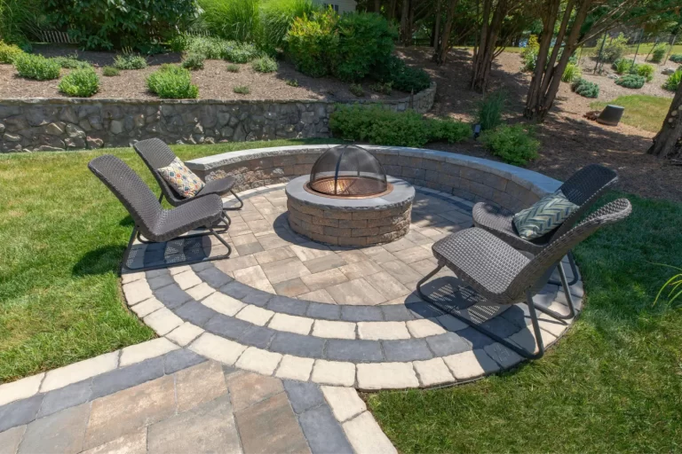 Circular stone patio with a fire pit and three wicker chairs with cushions in a landscaped backyard.