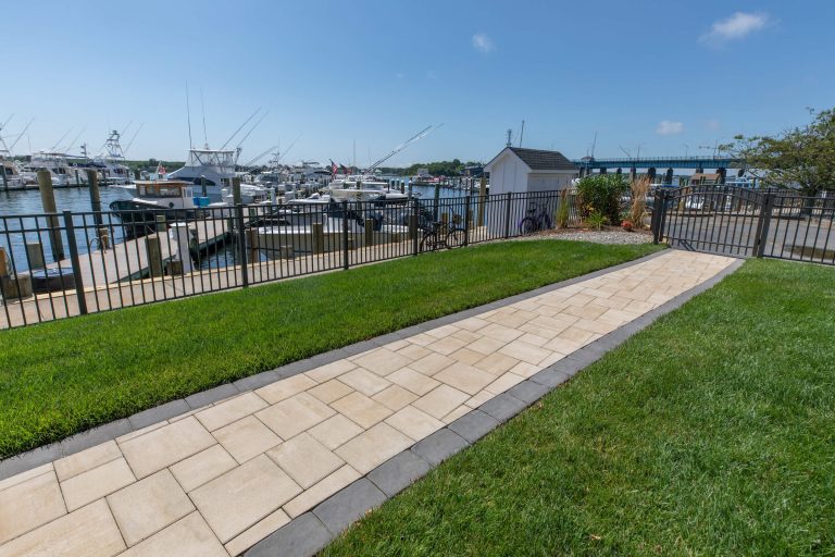 Paved walkway leads to a marina with boats docked, fenced grassy area, and a small white shed.