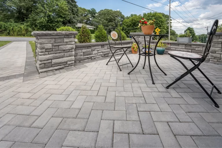Patio with gray pavers, a black metal table, two chairs, and potted flowers, bordered by a stone wall.