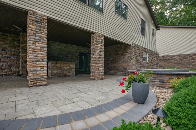 Stone patio with large pillars, potted red flowers, and landscaped garden next to a modern house.