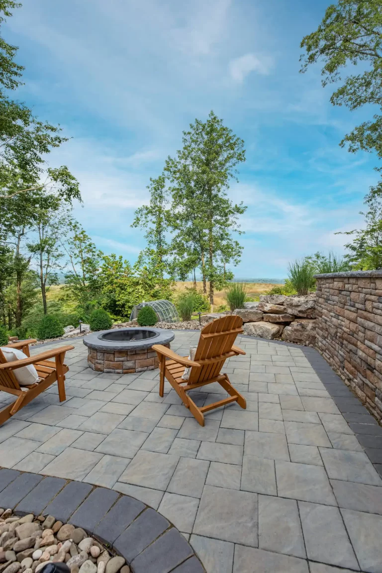 Two wooden chairs face a stone fire pit on a patio with a scenic view of trees and blue sky.