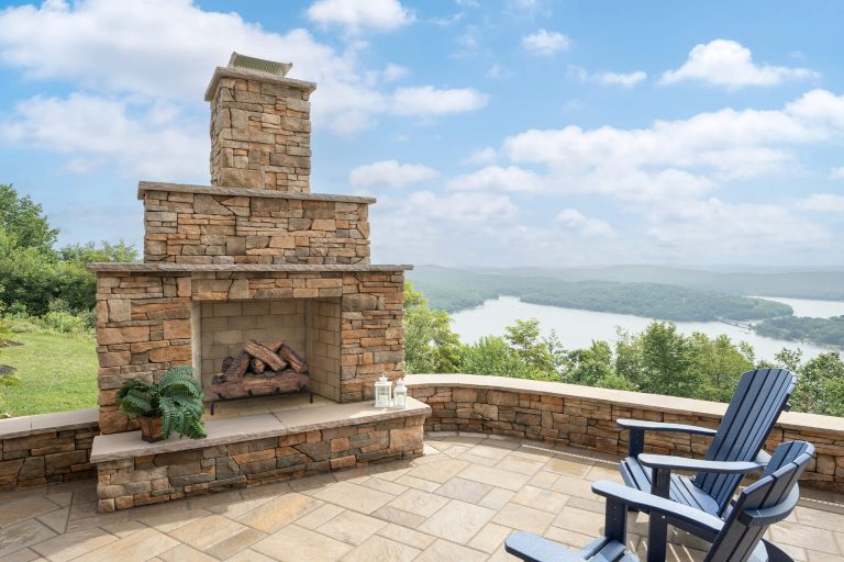 Stone outdoor fireplace and blue chairs on a patio overlooking a river and green hills under a partly cloudy sky.