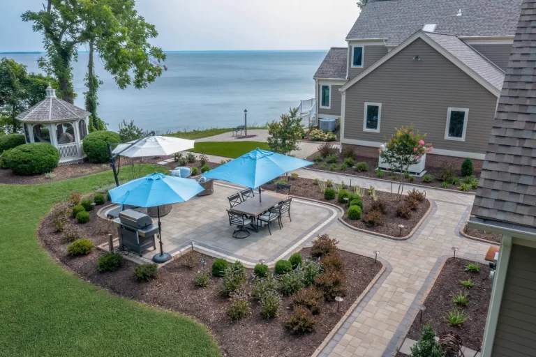 Backyard patio with blue umbrellas, seating, and grill overlooking a lake, next to a gazebo and landscaped garden.