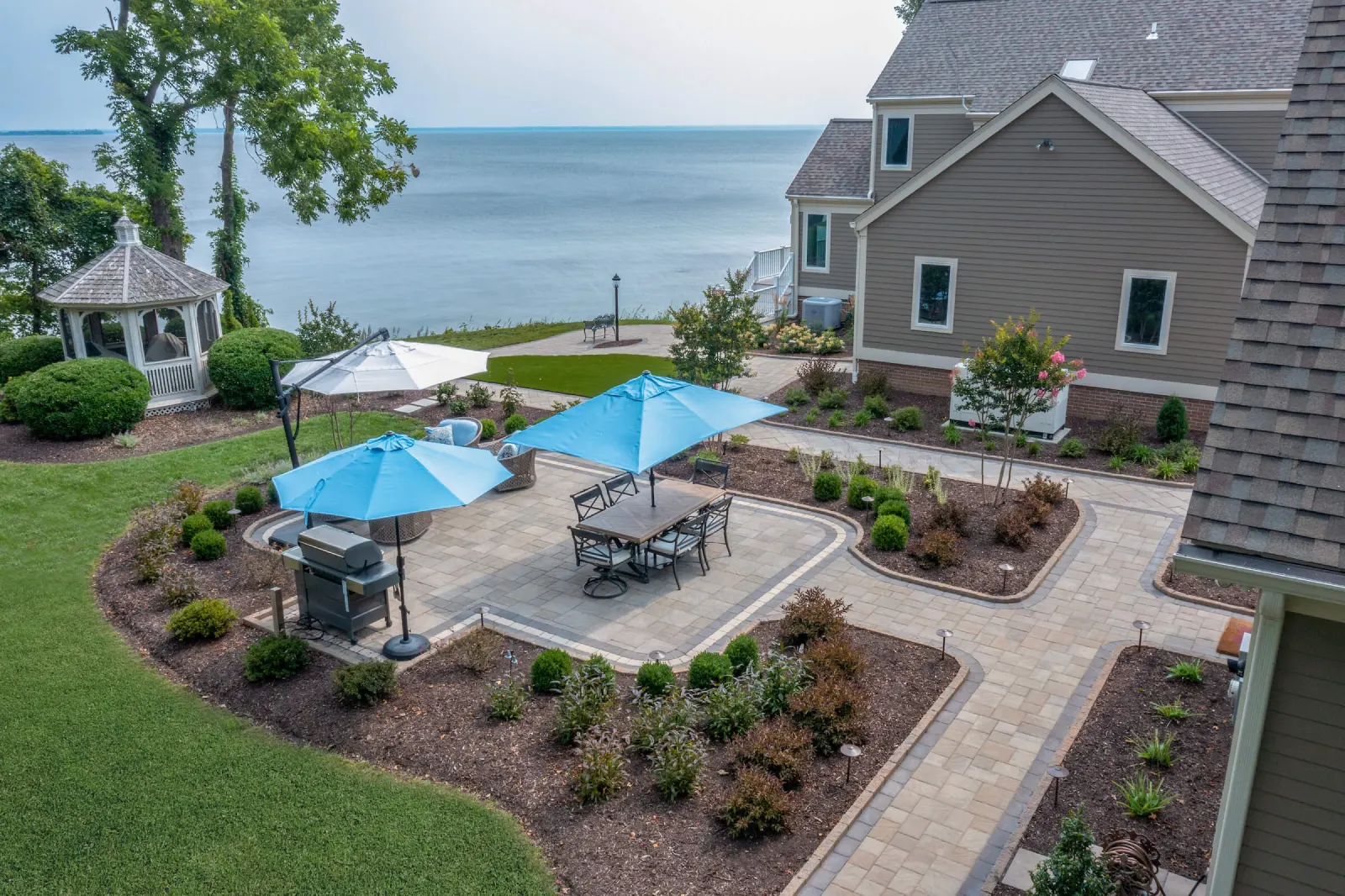 Backyard patio with blue umbrellas, seating, and grill overlooking a lake, next to a gazebo and landscaped garden.