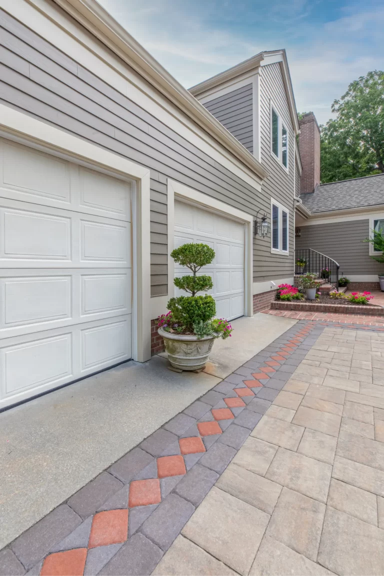 Two white garage doors on a house with gray siding, potted greenery, and a patterned stone driveway.