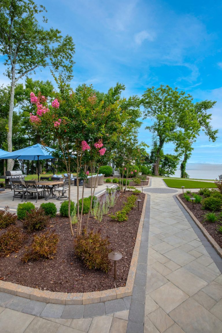 Stone walkway leads to a patio with seating, surrounded by trees, shrubs, and pink flowering plants.