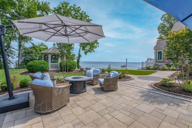 Patio with wicker chairs, a fire pit, and umbrella overlooking a lake, gazebo and trees in the background.