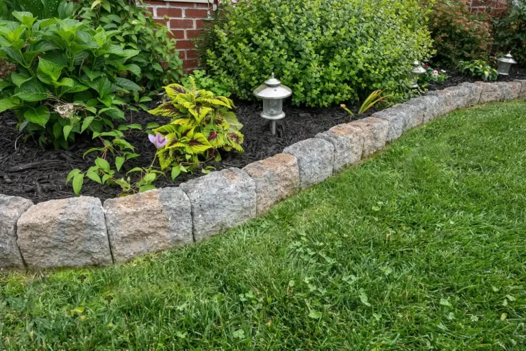 Stone edging separates a garden bed with green plants from a grassy lawn; lanterns are visible in the garden.