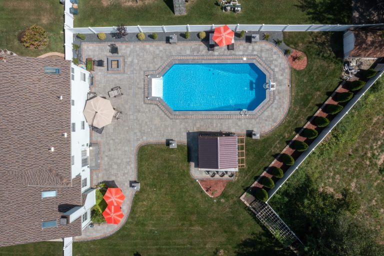 Aerial view of a backyard with a swimming pool, patio furniture, umbrellas, grass, and a white fence.