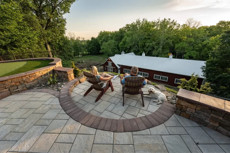 A couple and a dog relax on patio chairs overlooking a large red barn and lush green trees at sunset.