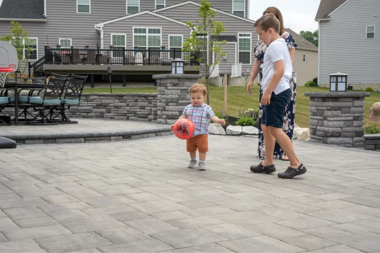 A toddler runs with a red ball while a woman and an older boy watch on a paved patio in a backyard.