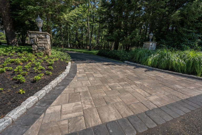 Stone paver driveway bordered by greenery and stone pillars with lanterns, surrounded by trees.