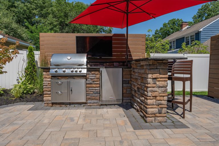 Outdoor kitchen with a grill, mini fridge, bar seating, and red umbrella on a stone patio.