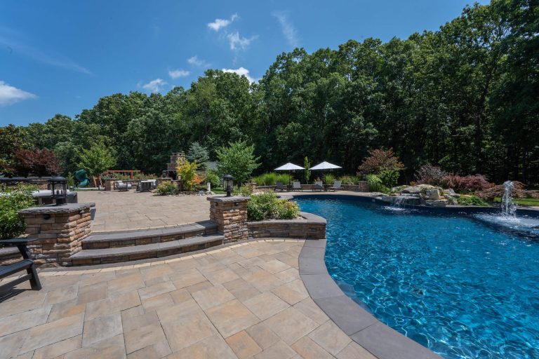 Spacious backyard patio with stone pavers, a pool, umbrellas, and lush green trees under a blue sky.
