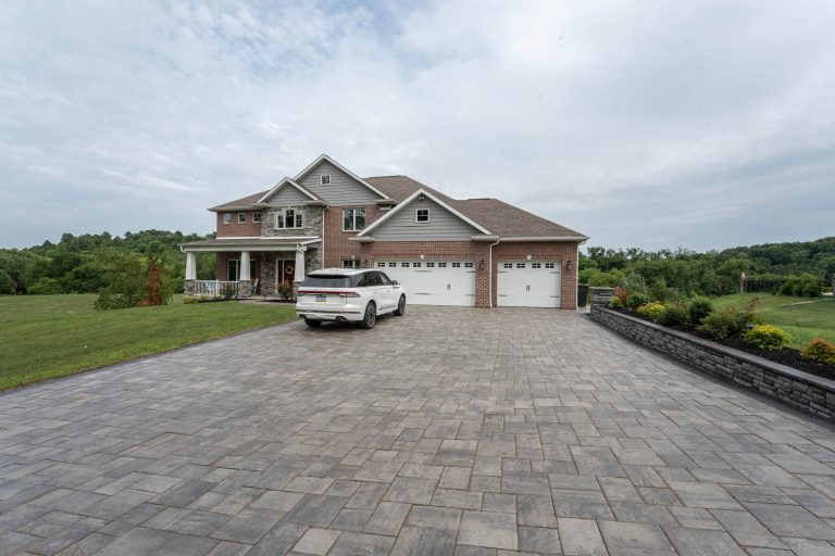 Large brick house with a three-car garage, white SUV parked on a wide stone driveway, surrounded by greenery.