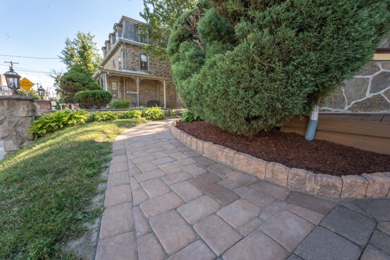Curved stone walkway beside a house, bordered by bushes, mulch, and a stone wall.