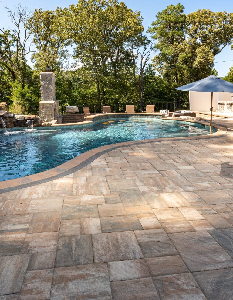 Curved pool with stone patio, lounge chairs, umbrella, and trees in the background on a sunny day.