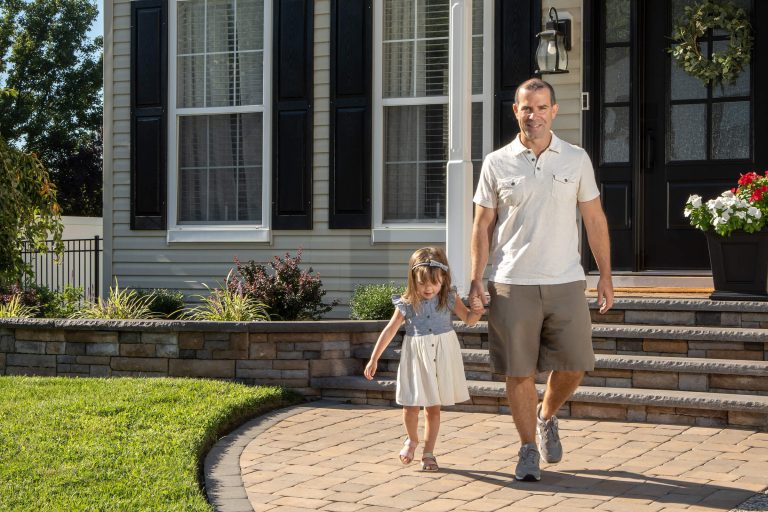 A man and young girl holding hands, walking on a stone pathway outside a house on a sunny day.