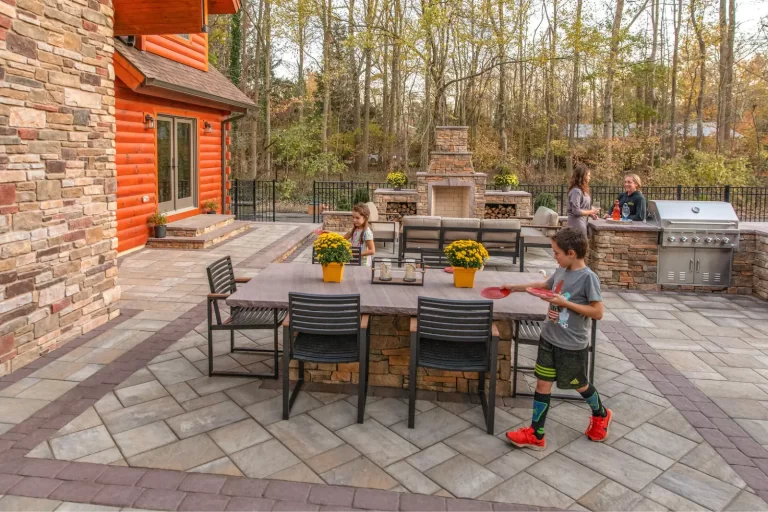 Children play near a patio table and grill as adults talk by a fireplace in a backyard with trees.
