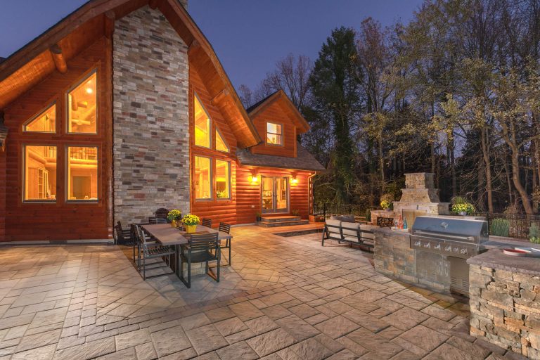 A warmly lit house with large windows, patio dining set, grill, and outdoor fireplace at dusk.