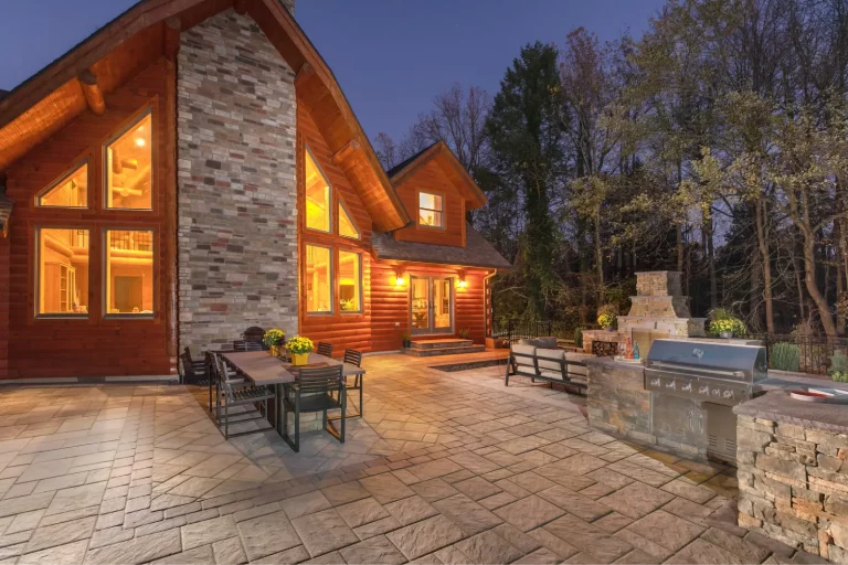 Large wooden house with warm lights, stone patio, outdoor dining area, and built-in grill at dusk.
