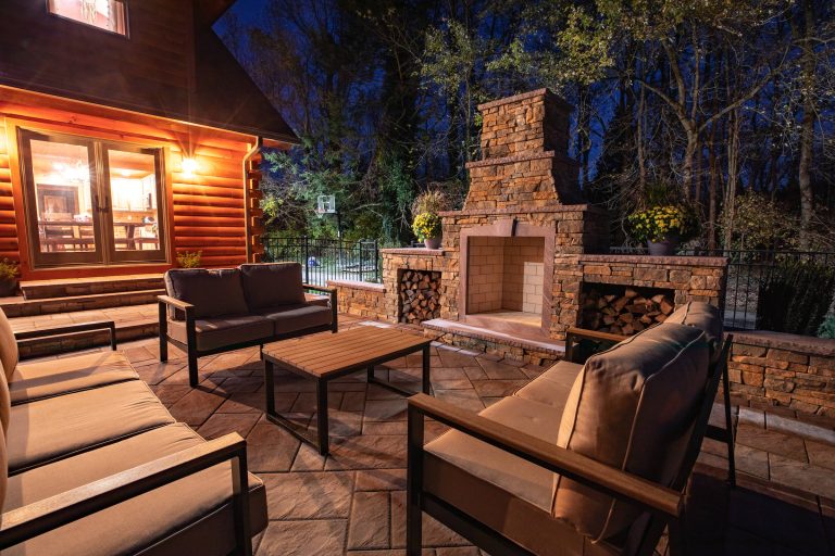 Cozy outdoor patio at night with cushioned seating, a wooden table, and a stone fireplace.