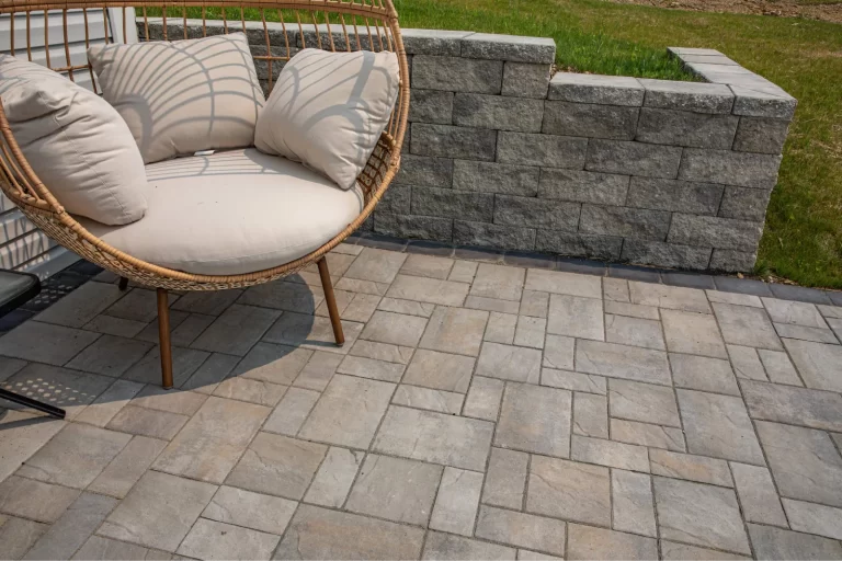 Round wicker chair with cream cushions on a stone patio beside a short stone retaining wall and grass.