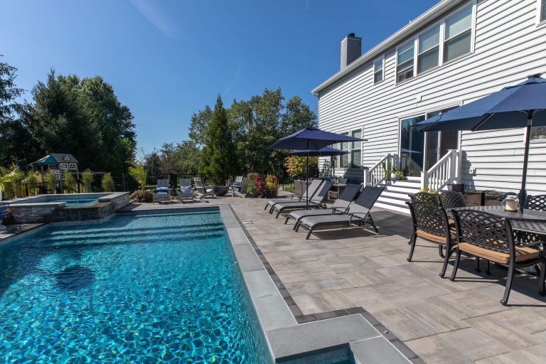 Backyard with a pool, lounge chairs, patio table, umbrellas, and a white house on a sunny day.