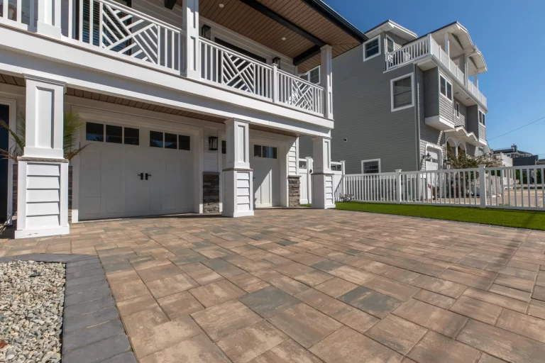 Modern house with white garage doors, paved driveway, white fence, and green lawn under a blue sky.