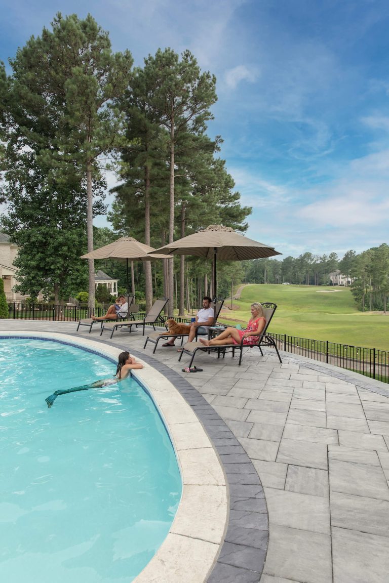 Three people relax on lounge chairs by a pool while one person swims nearby; trees and grass in the background.