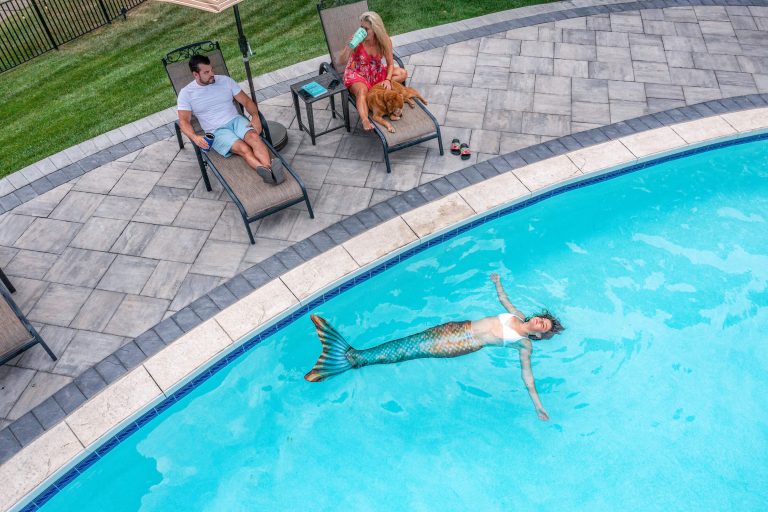 A person in a mermaid tail floats in a pool while two people relax on lounge chairs nearby.