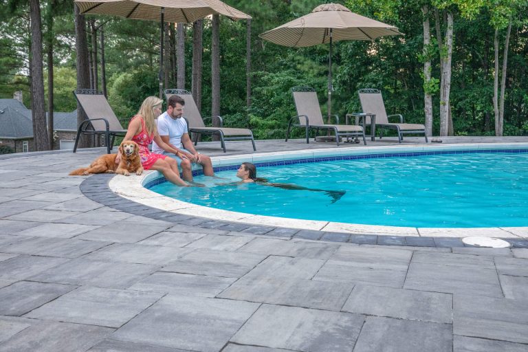 A couple and their dog sit by a pool while a person in a mermaid tail swims nearby.