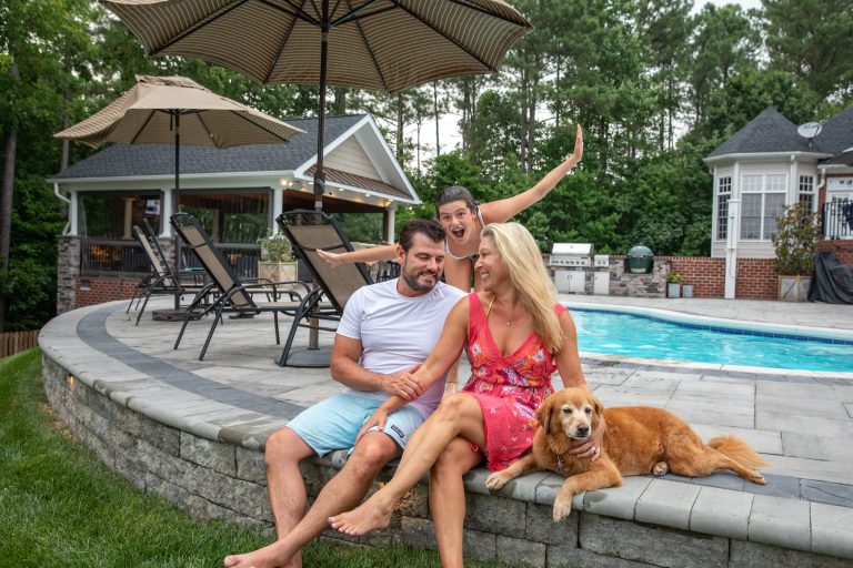 A couple sits by a pool with a dog, while a smiling woman photobombs them in the background.