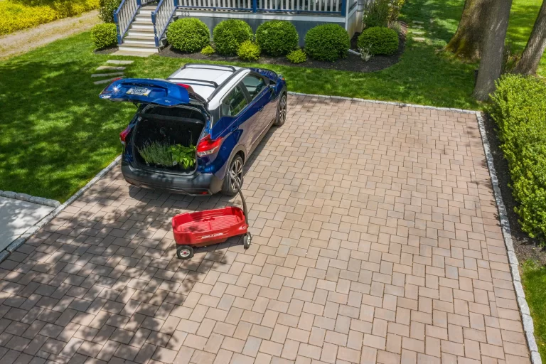 Blue SUV with open trunk and plants inside, next to a red wagon on a brick driveway near a house.
