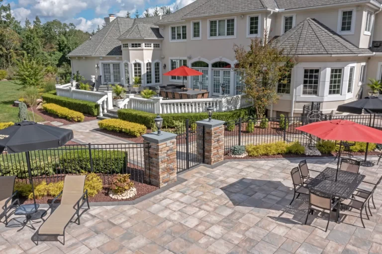 Large backyard patio with lounge chairs, outdoor dining tables, umbrellas, and landscaped garden by a big house.
