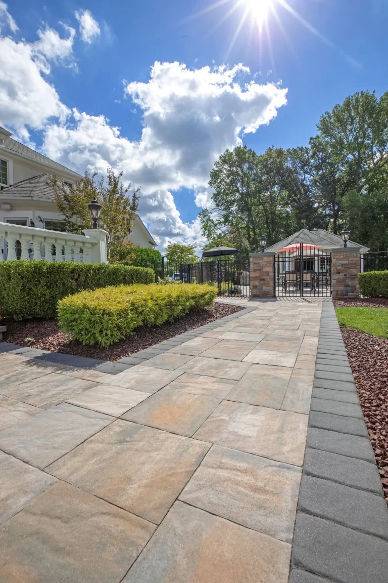 Wide stone walkway leading to a gated entrance, with bushes, a house, and a bright sunny sky.