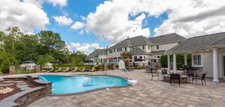 Luxury pool with lounge chairs, patio tables, and a large house in the background under a partly cloudy sky.
