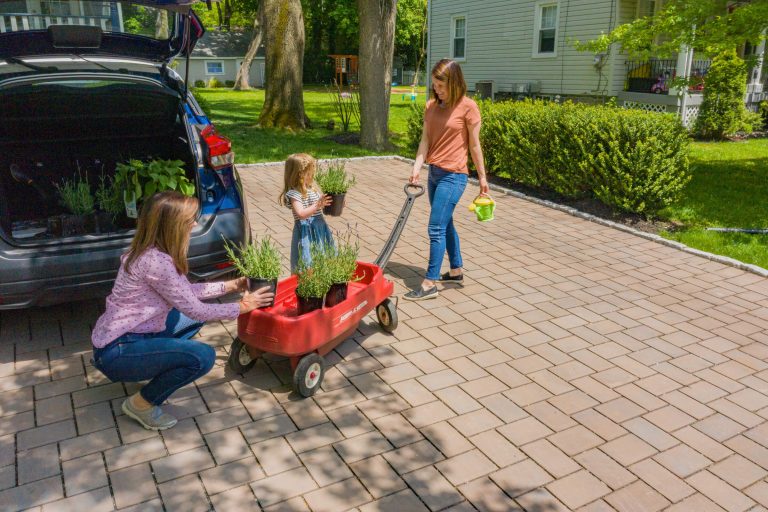 Two women and a girl unload plants from a car into a red wagon on a sunny driveway.