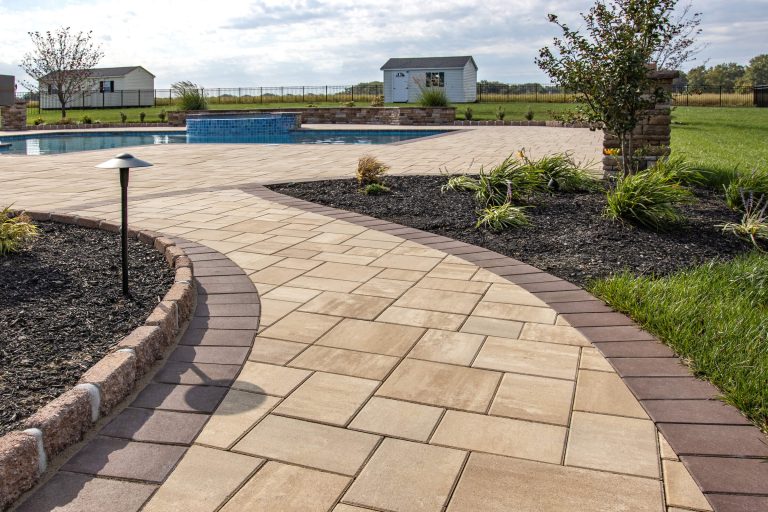 Curved stone pathway leads to a patio and pool, surrounded by grass, plants, and landscaped beds.