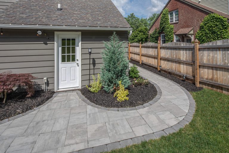 Curved stone walkway with plants beside a gray house and wooden fence on a sunny day.