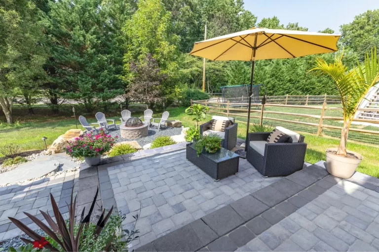 Patio with wicker chairs, yellow umbrella, fire pit, and lawn chairs, surrounded by trees and a wooden fence.