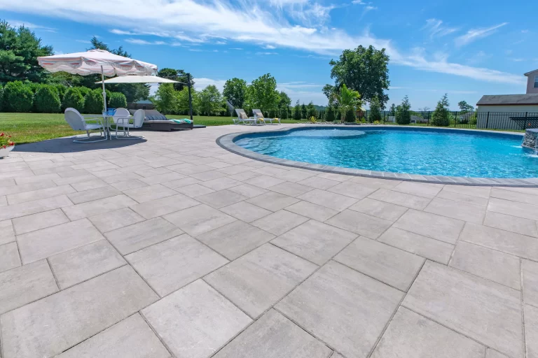 Sunny backyard pool with lounge chairs, umbrellas, and large stone patio surrounded by grass and trees.