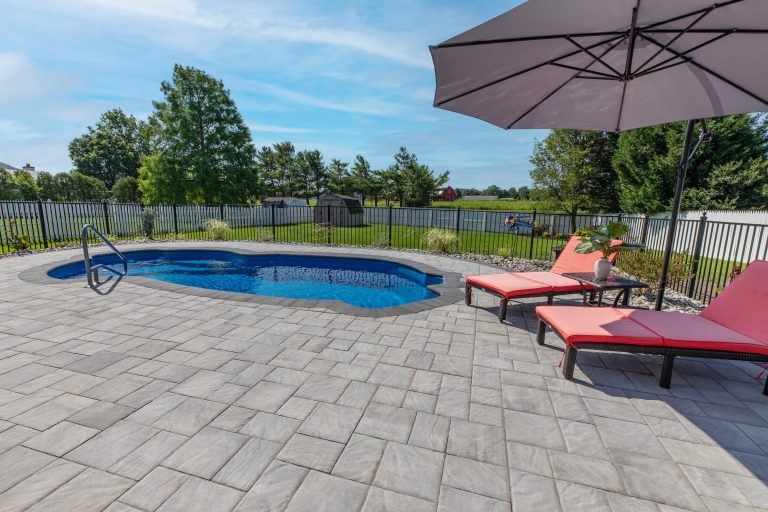 Small backyard pool with metal rail, pink lounge chairs, umbrella, and a view of grass and trees beyond a fence.