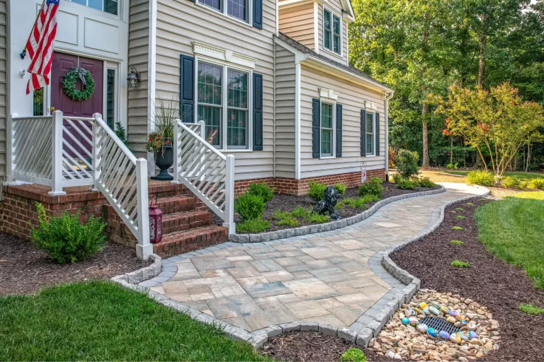 Curved stone walkway leads to a beige house with a brick porch and landscaped garden beds.