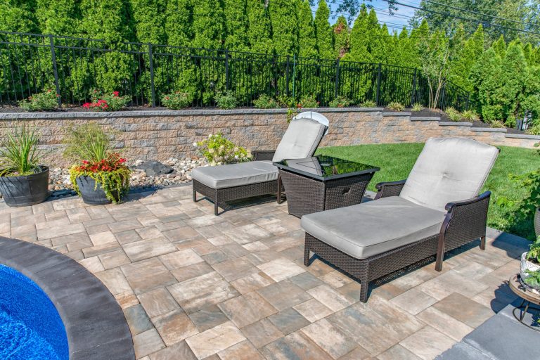 Two lounge chairs and a small table sit on a stone patio next to a pool, with greenery in the background.