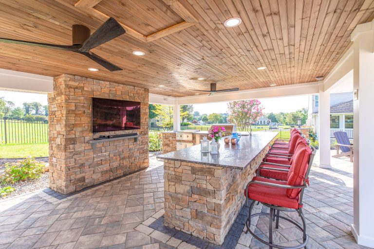 Covered outdoor kitchen with stone bar, red barstools, ceiling fan, TV, and garden view.